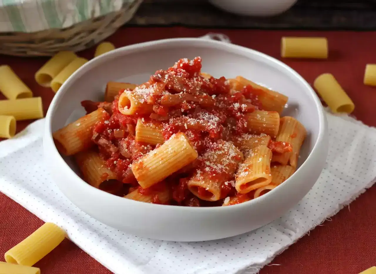 En skål med rig pasta amatriciana, toppad med riven ost. Omgiven av okokt pasta.