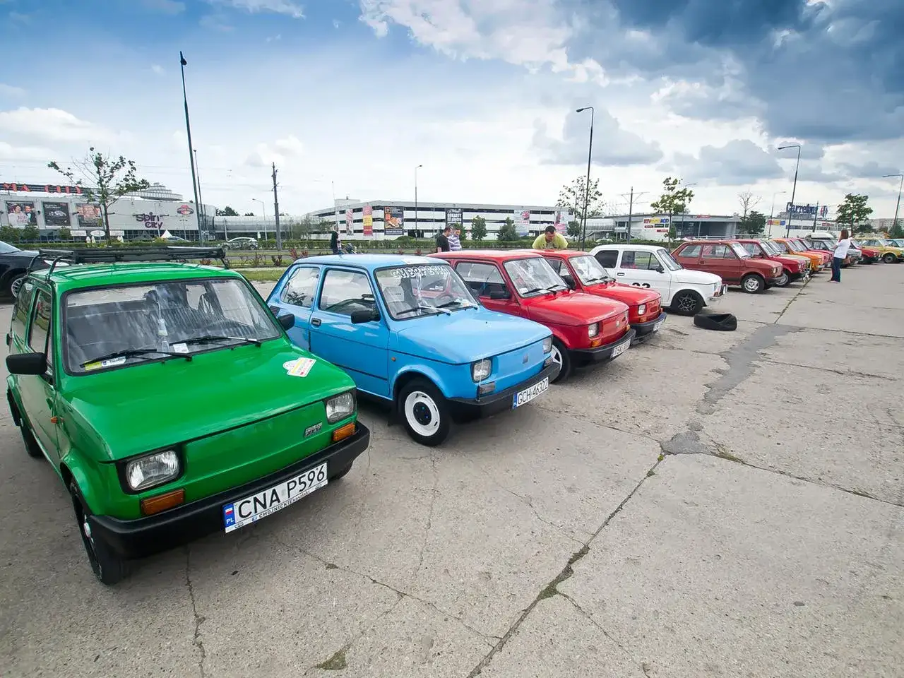 Rząd kolorowych **polskich fiatów 126p** zaparkowanych na placu. Zielony maluch z przodu, za nim niebieski, czerwone i białe.