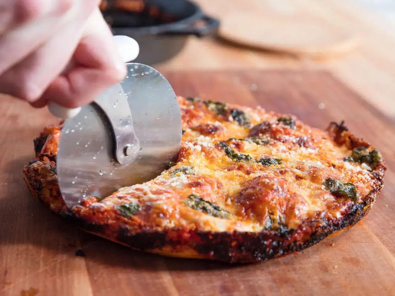 Une main coupe une pizza &agrave; la po&ecirc;le dor&eacute;e et garnie de fromage fondant et d'&eacute;pinards sur une planche en bois.