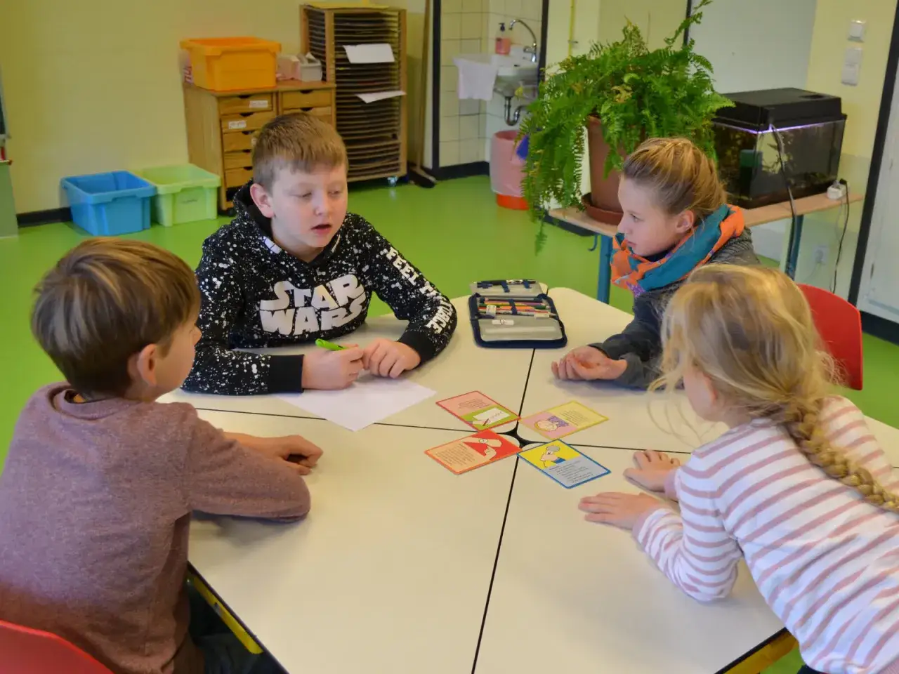 Vier Kinder sitzen an gruppentische grundschule und spielen mit Karten. Ein Junge mit Star Wars Hoodie erkl&auml;rt etwas.