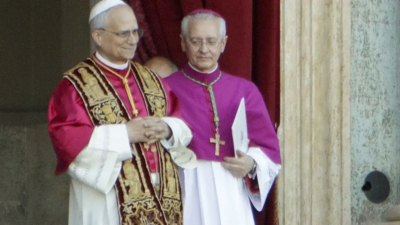 Dwóch duchownych w bogato zdobionych szatach. Jeden z nich, prefekt, uśmiecha się, trzymając dłonie złożone do modlitwy.