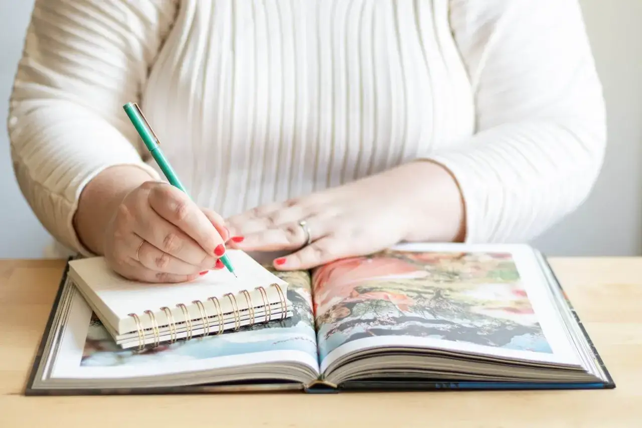 Frau schreibt Notizen in ein Notizbuch, um ein **Jahrbuch erstellen** zu können. Ein aufgeschlagenes Fotoalbum liegt daneben.