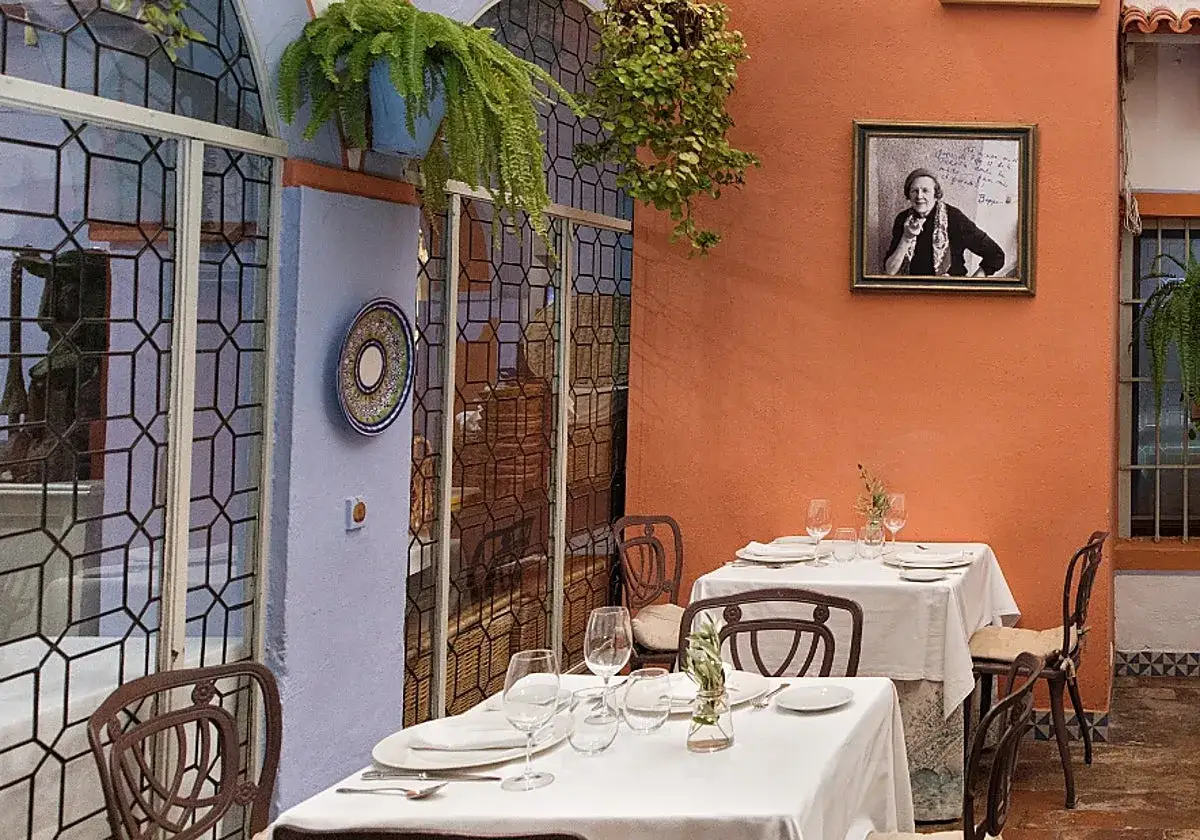 Mesas puestas en un patio andaluz, ideal para saber donde comer en C&oacute;rdoba. Un retrato adorna la pared naranja.