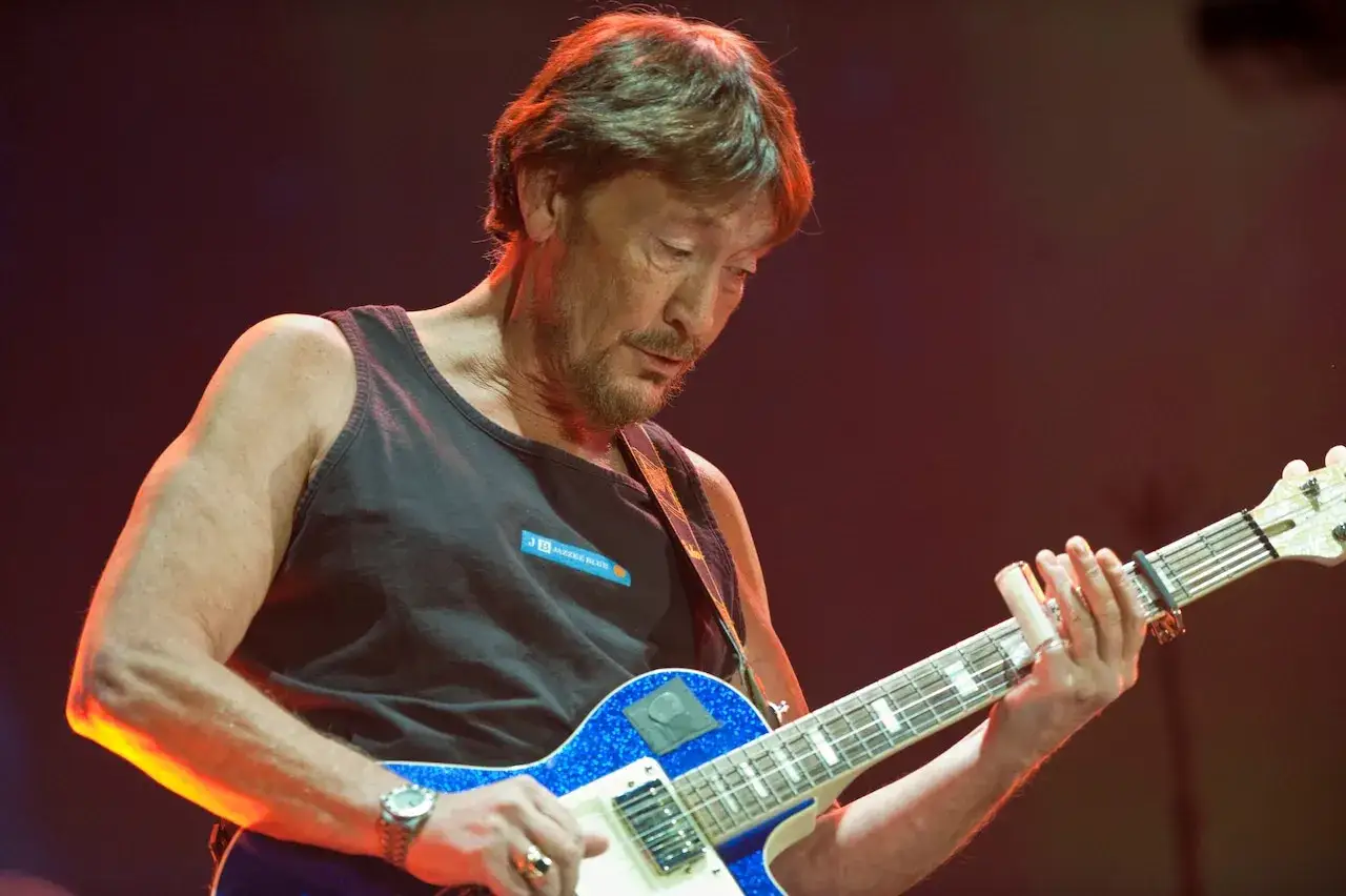 Chris Rea, w czarnej koszulce bez rękawów, gra na błyszczącej, niebieskiej gitarze elektrycznej.