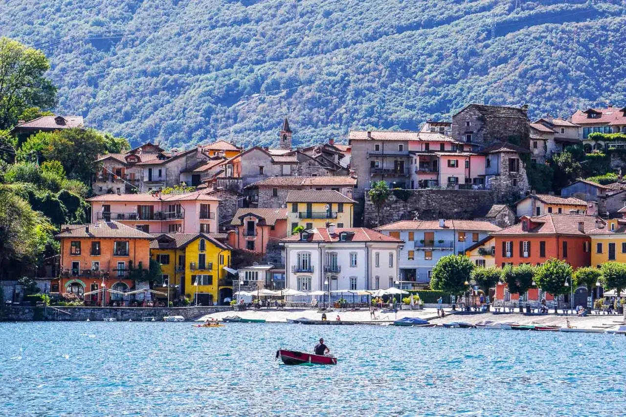 Un uomo in barca sul lago, con case colorate che si arrampicano sulla collina. Questo &egrave; un scorcio di dove si trova Domodossola, un luogo incantevole.