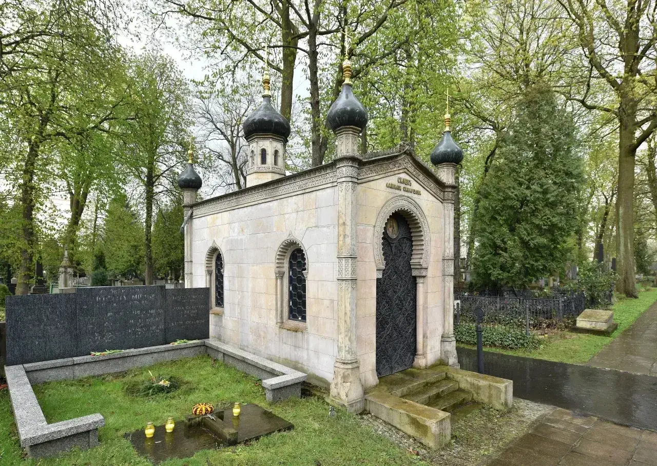 Imponujący, murowany grób z kopułami i ozdobnymi detalami, otoczony zielenią cmentarza.