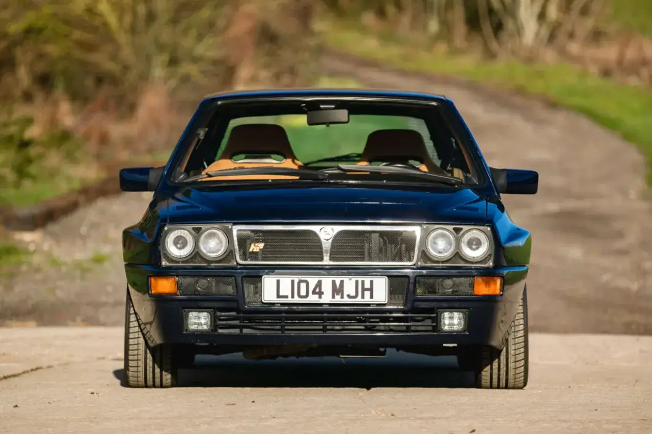 Ikoniczna Lancia Delta Integrale w ciemnoniebieskim kolorze, z brązowymi siedzeniami, gotowa na rajdową przygodę.
