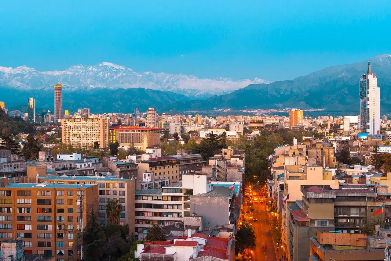 Widok na rozświetlone miasto Santiago de Chile o zmierzchu, z ośnieżonymi Andami w tle.