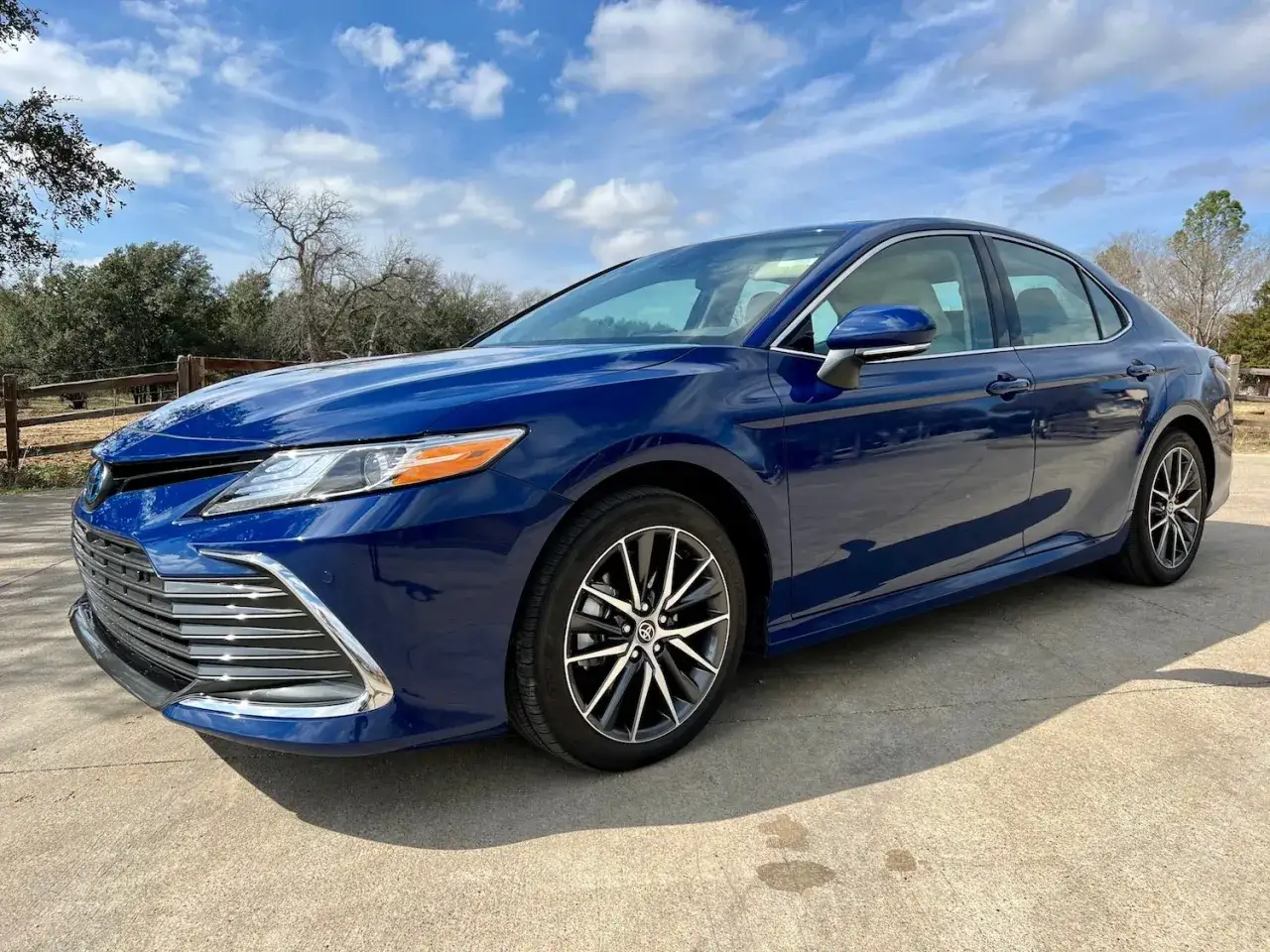 Błękitna Toyota Camry 2023 zaparkowana na podjeździe, z drzewami i niebieskim niebem w tle.