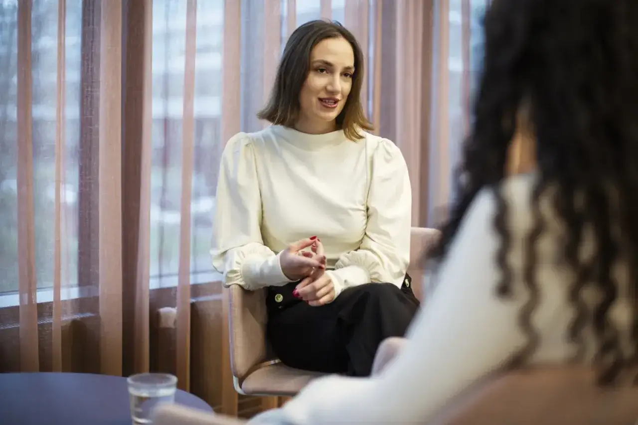 En kvinna med kort brunt h&aring;r och vit blus sitter i en stol och pratar. Hon verkar f&ouml;rklara psykolog utbildning krav.