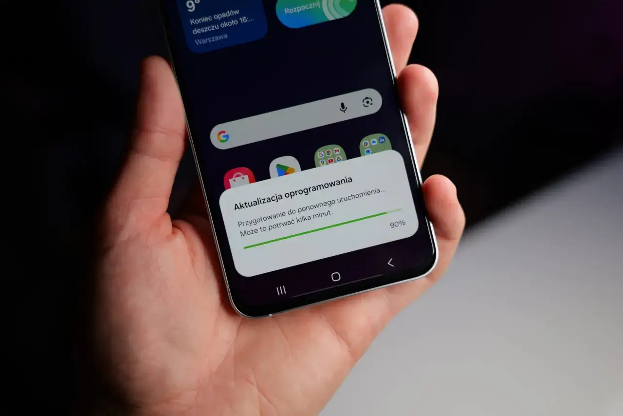 Dłoń trzyma smartfon z powiadomieniem o aktualizacji oprogramowania do android 14. Proces zakończony w 90%.