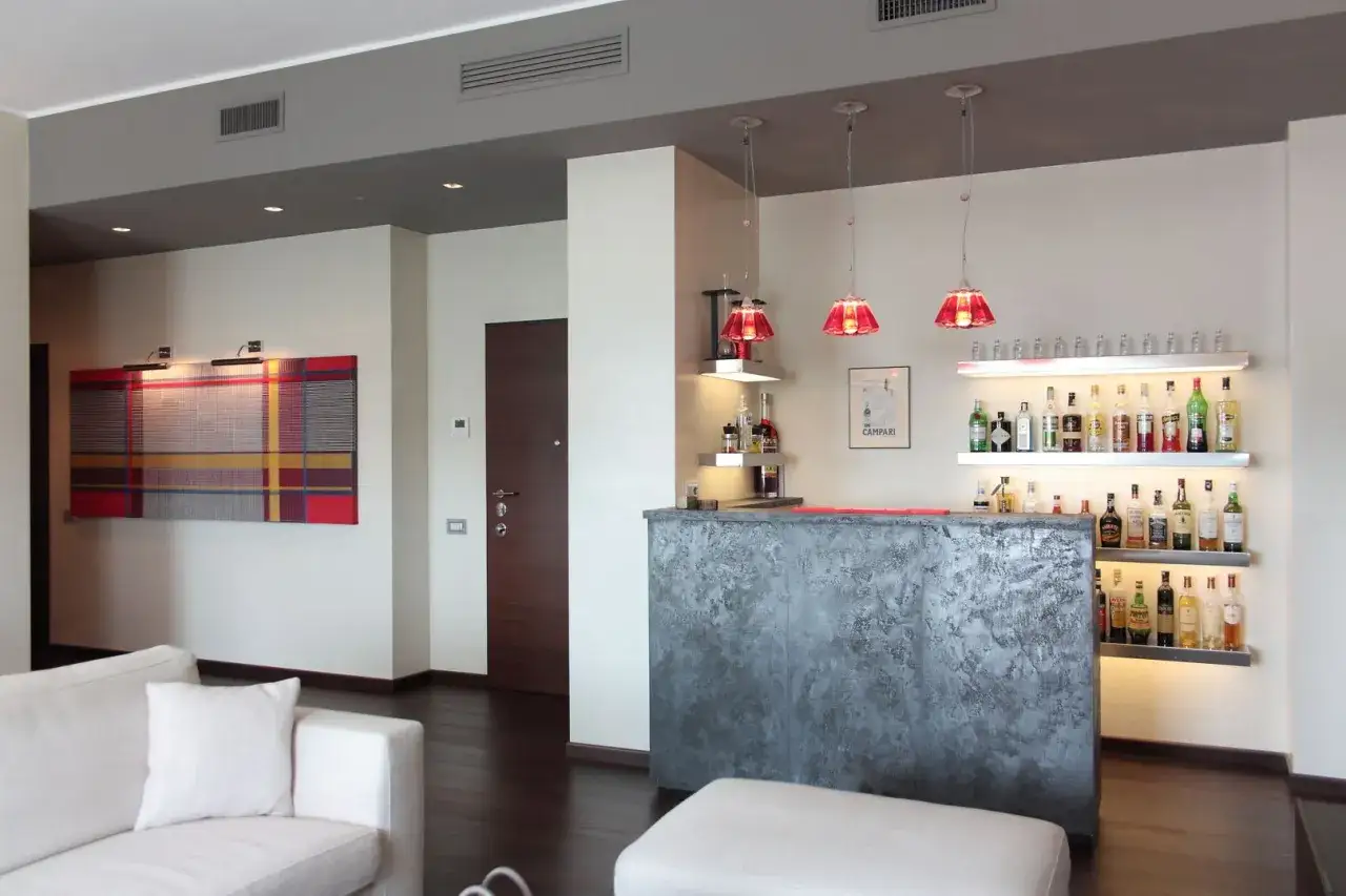 Elegante angolo bar con bancone grigio materico, bottiglie di liquori su mensole illuminate e lampade rosse.