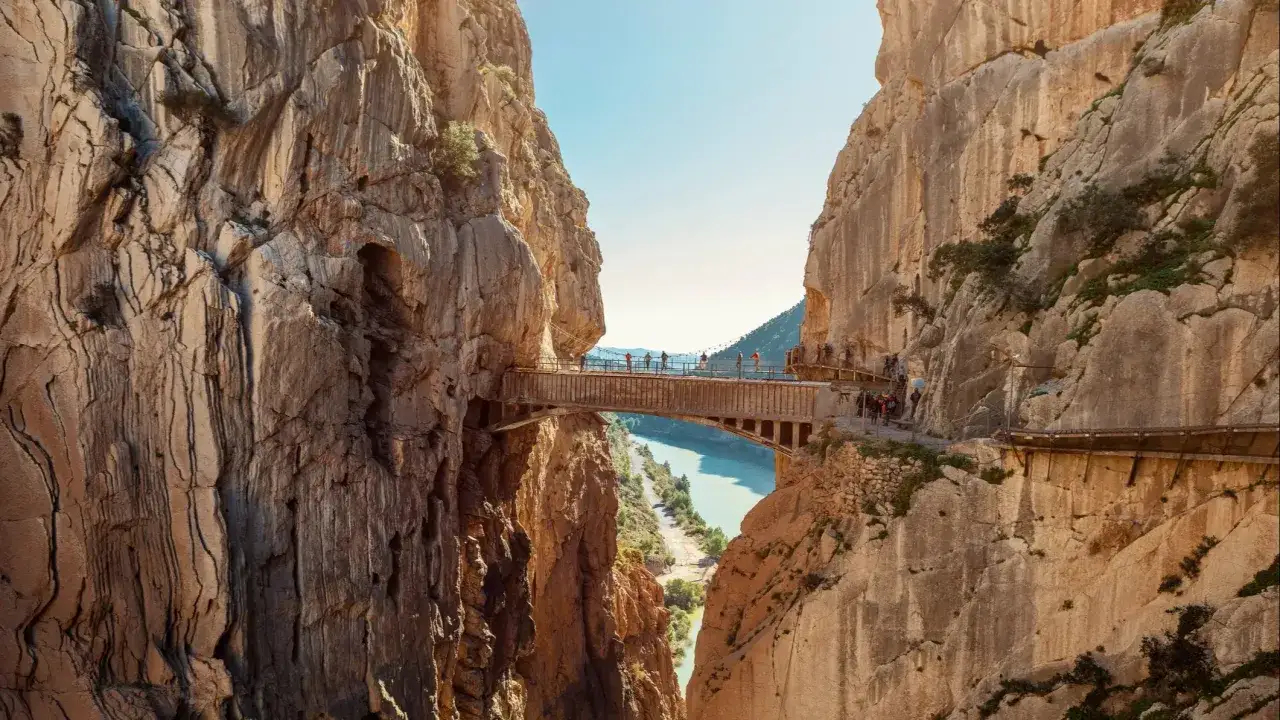 Ein atemberaubender tagesausflug malaga: Menschen &uuml;berqueren eine Br&uuml;cke in einer tiefen Schlucht, umgeben von steilen Felsw&auml;nden und t&uuml;rkisfarbenem Wasser.