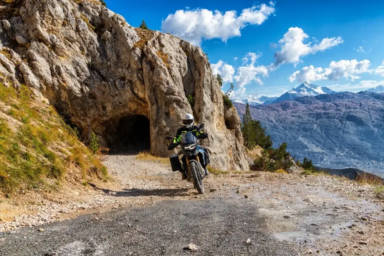 Un motociclista esplora un sentiero di montagna vicino a un tunnel roccioso. La **Jafferau moto** affronta il terreno accidentato con uno sfondo di maestose vette innevate.
