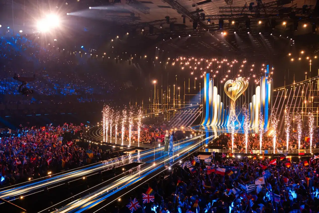Publiken jublar med flaggor under en glittrande scen. En artist står på scenen, redo för Eurovision Song Contest startordning.