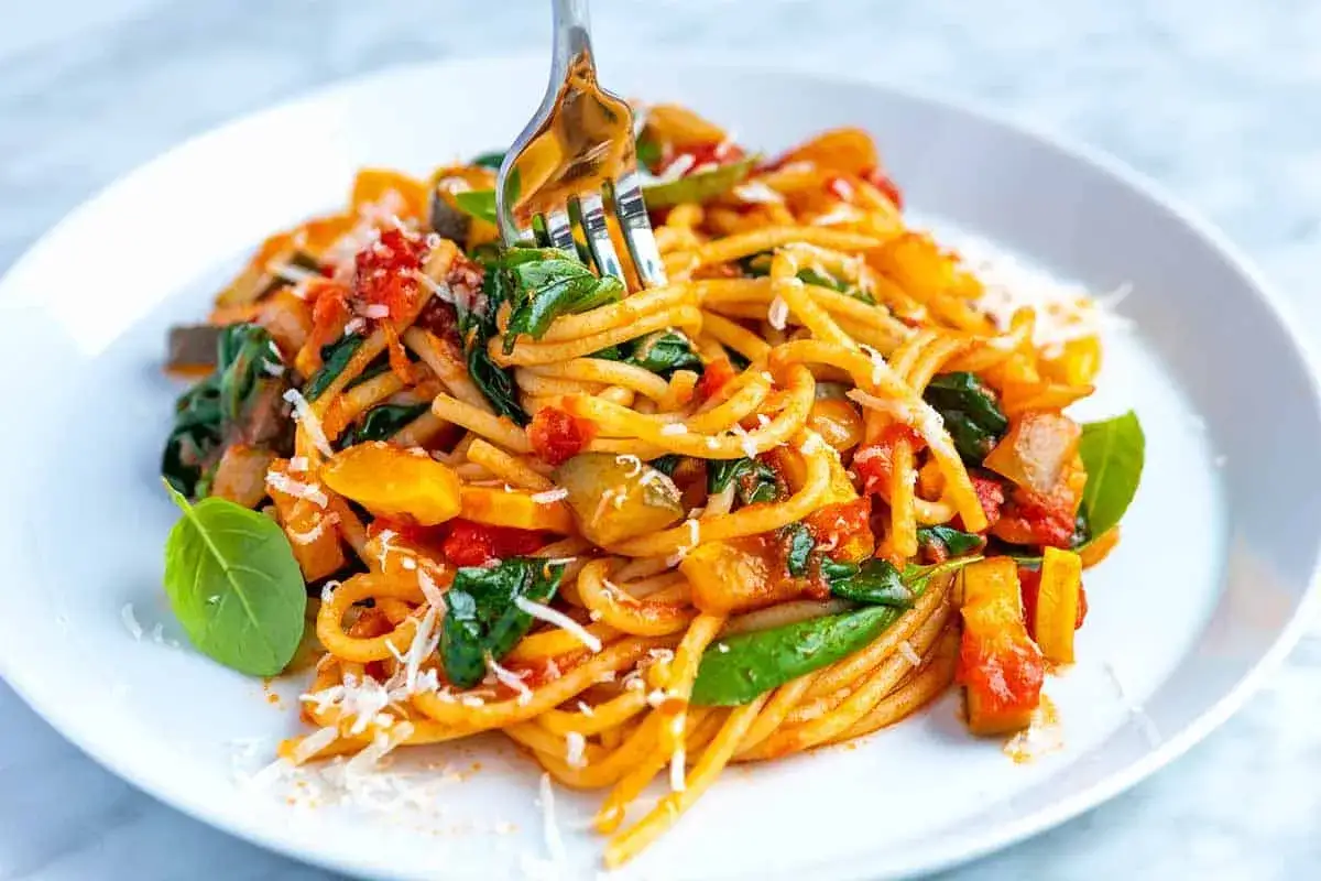 Un tenedor recoge un plato de pasta con verduras, con trozos de calabacín, tomate y espinacas, espolvoreado con queso rallado.