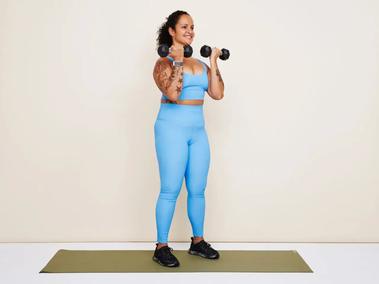 Femme souriante faisant de la musculation à la maison avec des haltères, portant un ensemble bleu vif.