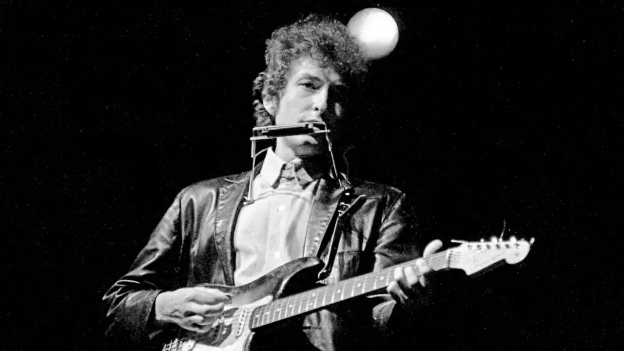 Bob Dylan gra na gitarze i harmonijce, tworząc swoje legendarne utwory.