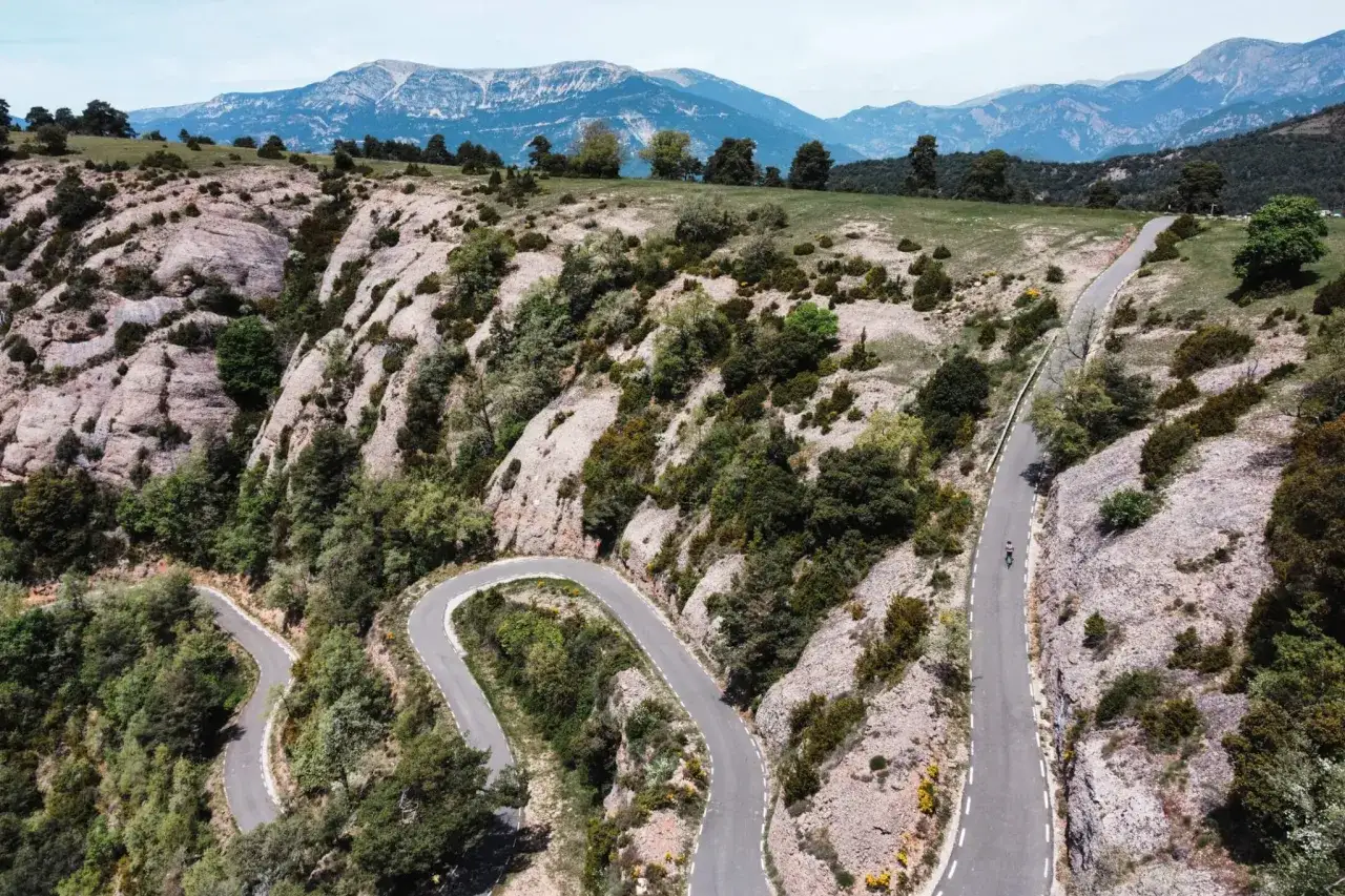 Carretera serpenteante en la Serra de Busa, con un ciclista solitario ascendiendo entre rocas y vegetaci&oacute;n.