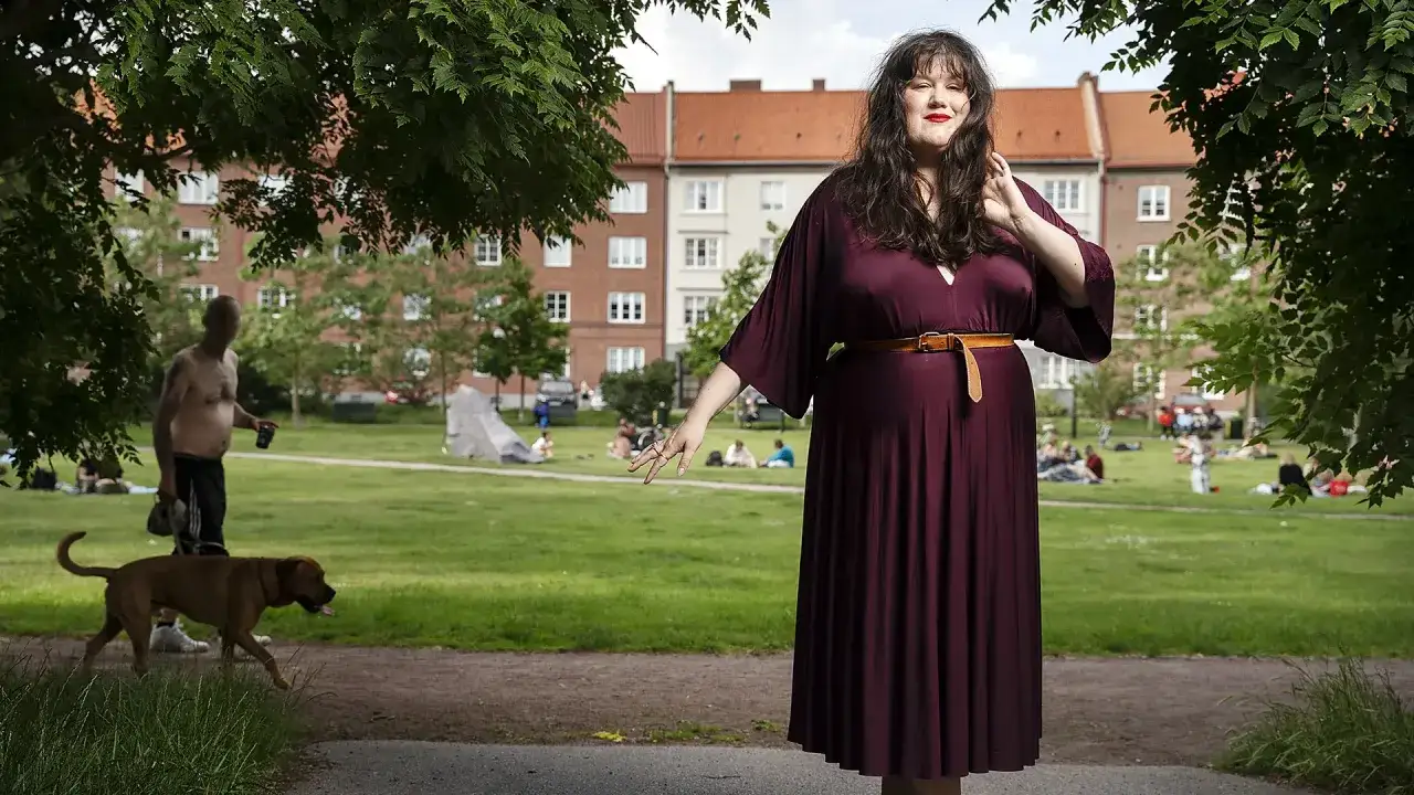 Maria Maunsbach i en vinröd klänning poserar i en park. I bakgrunden syns fler människor och bostadshus.