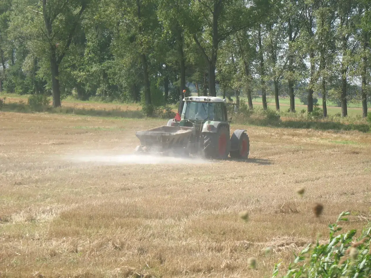 Traktor rozsypuje wapno węglanowe na polu, tworząc chmurę pyłu.