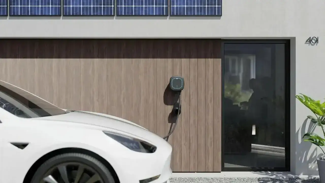 Biały samochód elektryczny podłączony do ładowarki przy nowoczesnym domu z panelami słonecznymi. Zastanawiasz się, ile kosztuje naładowanie samochodu elektrycznego?