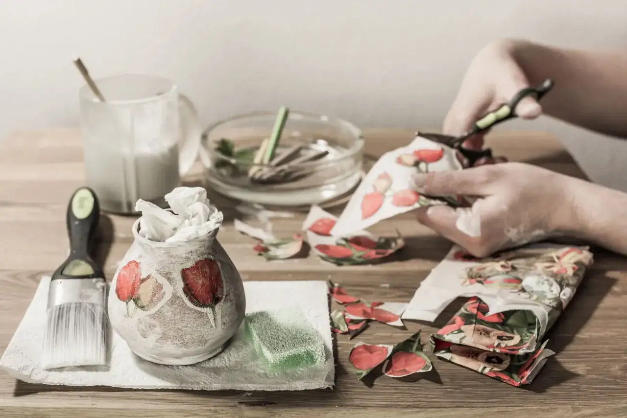 Ręce wycinają wzory z serwetek do decoupage. Wazonik ozdobiony kwiatami, pędzel i klej czekają na dalsze prace.