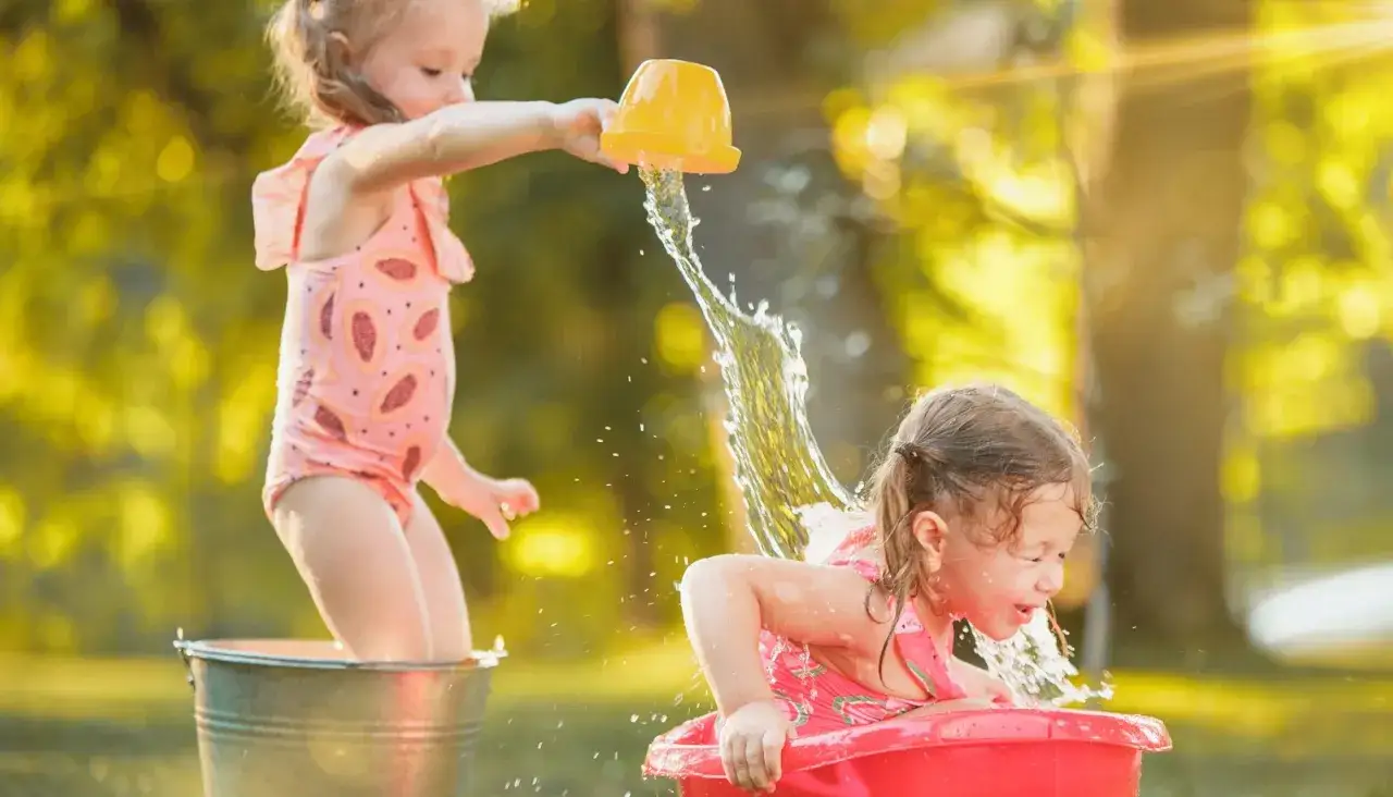 Bambine felici si divertono con giochi d'acqua sotto il sole, una versa acqua sull'altra con un secchiello giallo.