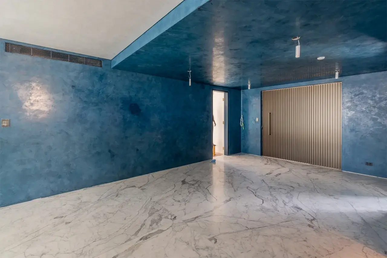 Habitaci&oacute;n con paredes y techo de estuco azul vibrante, suelo de m&aacute;rmol blanco y una puerta de madera acanalada.