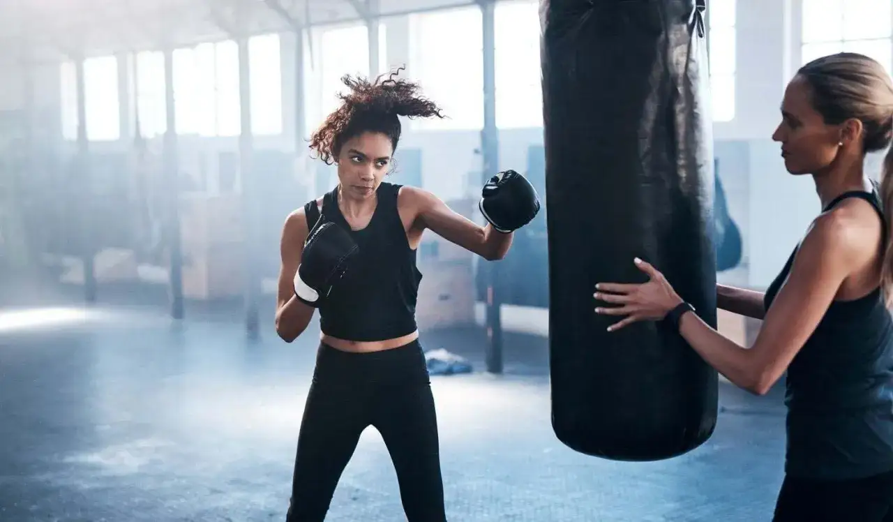 A woman in boxing gloves throws a punch at a heavy bag, her trainer holding it steady. This is what boxing everyday looks like.