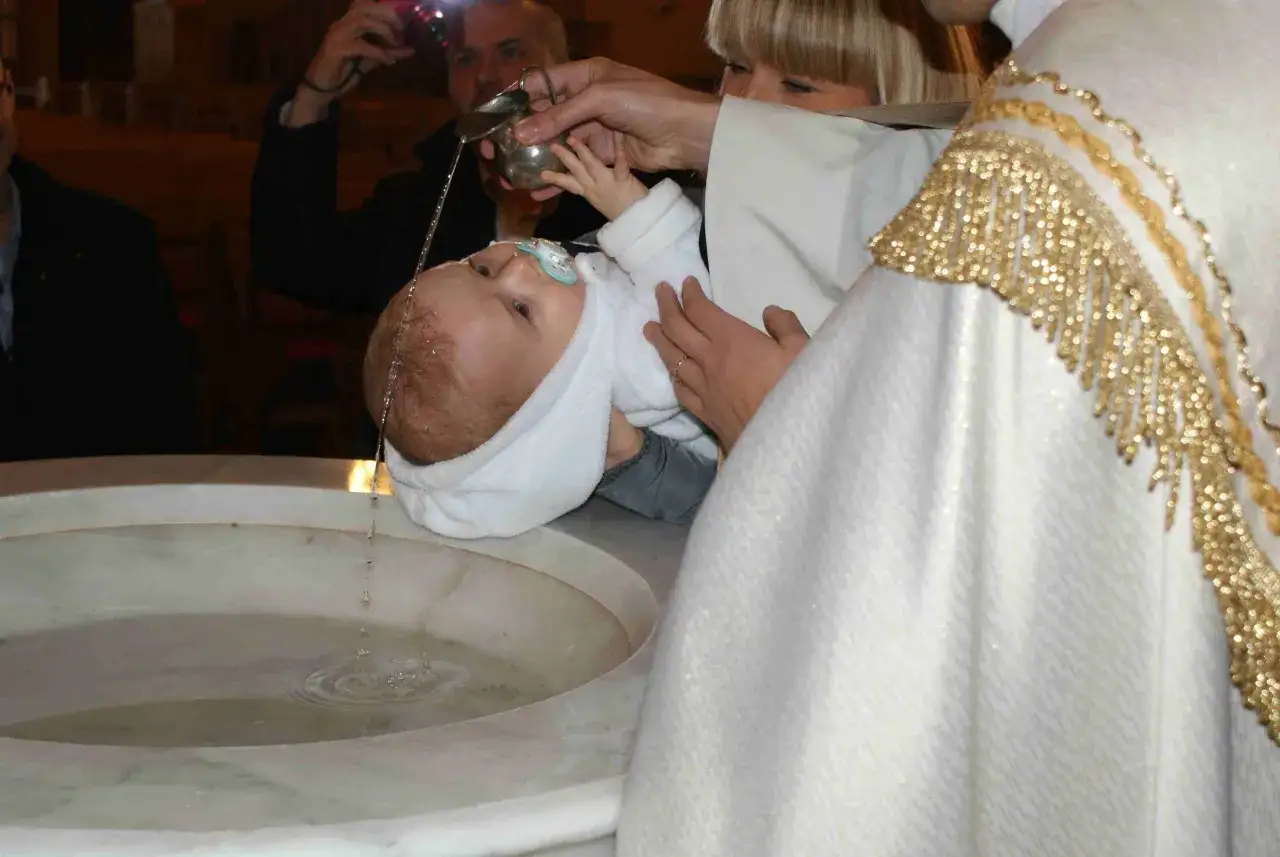 Chrzest dziecka bez mszy. Kapłan polewa gł&oacute;wkę niemowlęcia wodą ze srebrnej chrzcielnicy.