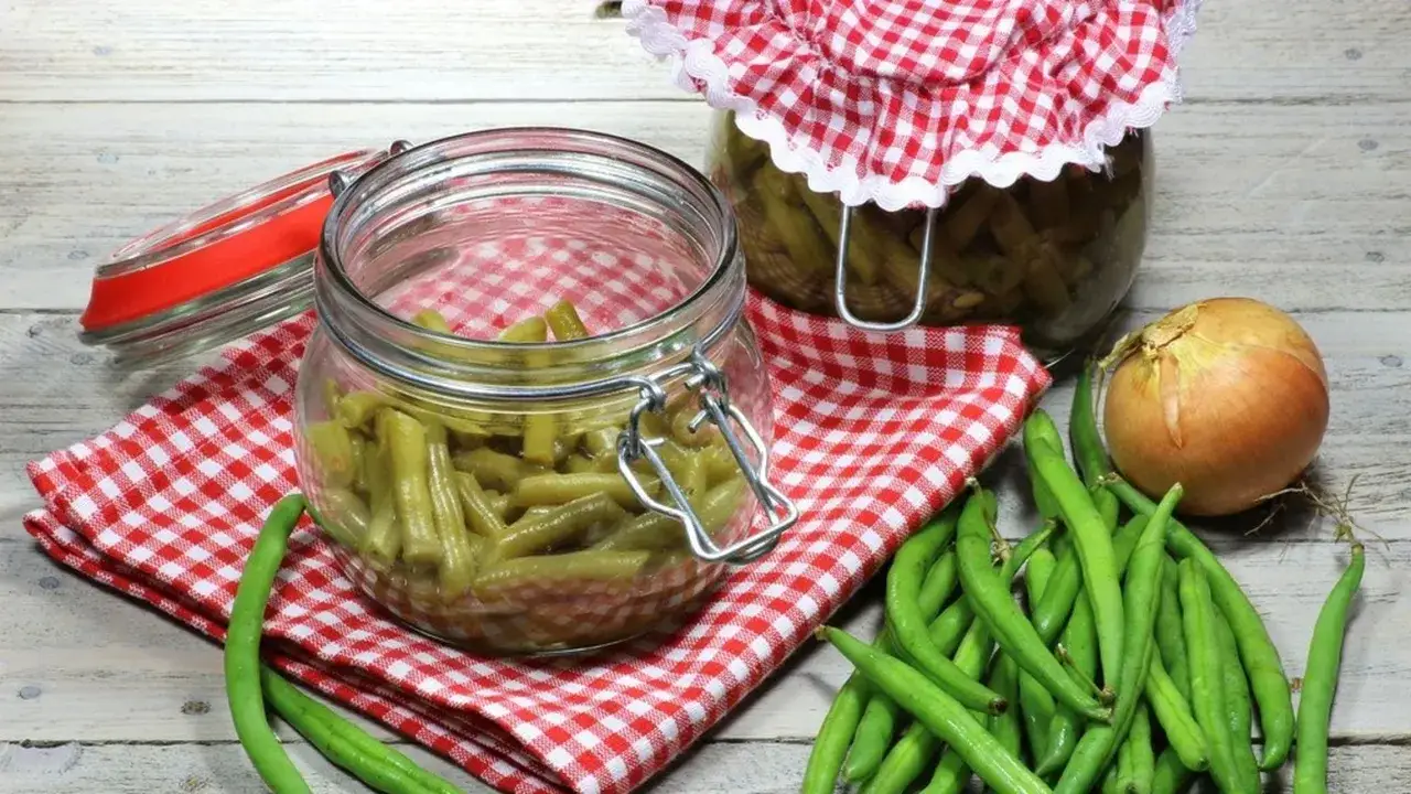 Fagiolini sott'olio in agrodolce, pronti per essere gustati. Un barattolo aperto e uno chiuso, con fagiolini freschi e una cipolla a lato.
