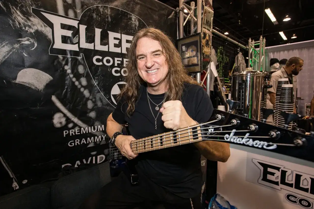 Dave Ellefson, muzyk z długimi włosami, trzyma gitarę basową Jackson. W tle logo Ellefson Coffee Co.