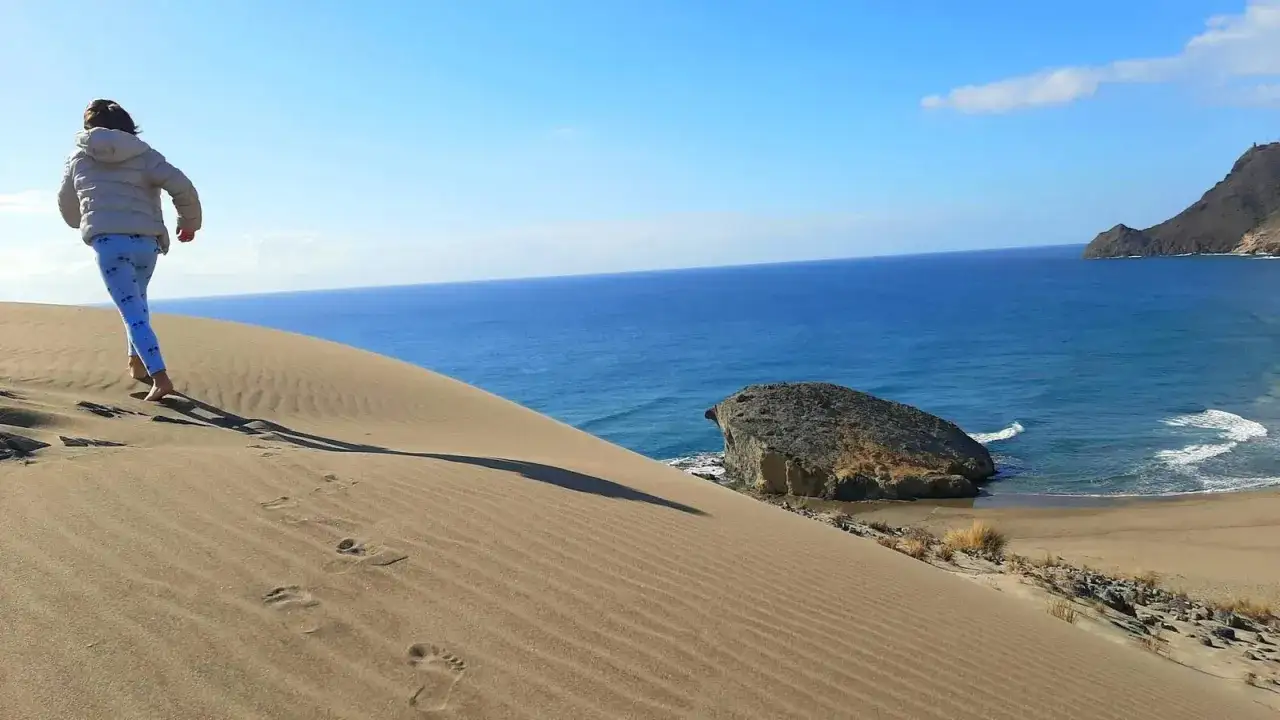 Niña corre descalza por la duna de Monsul, dejando huellas en la arena dorada. El mar azul intenso y una roca solitaria completan la escena.