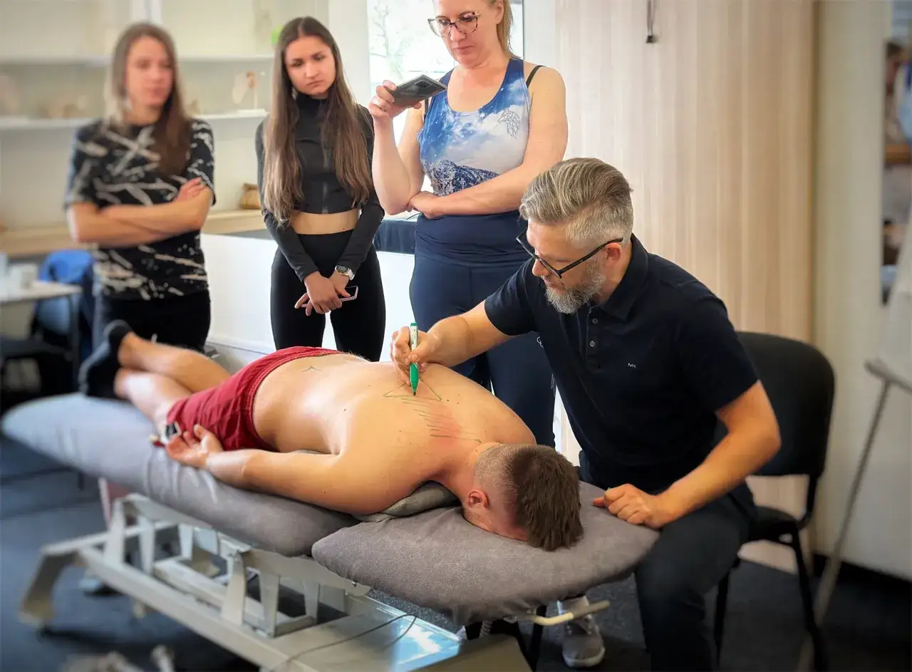 Terapeuta zaznacza punkty na plecach pacjenta, przygotowując się do zabiegu pinopresury. Obok stoją obserwatorzy.