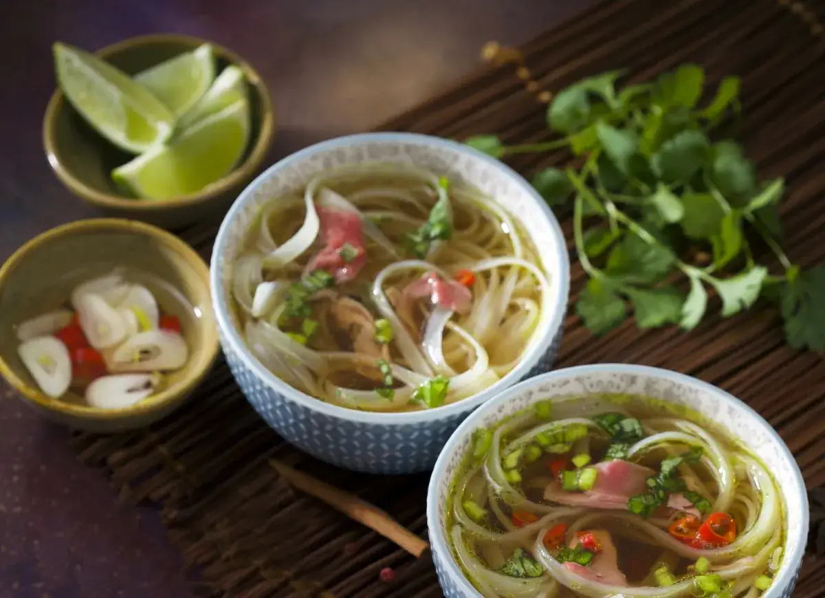 Dwie miseczki aromatycznej zupy pho bo z wołowiną, makaronem ryżowym i świeżą kolendrą. Obok limonka i marynowany czosnek z chili.