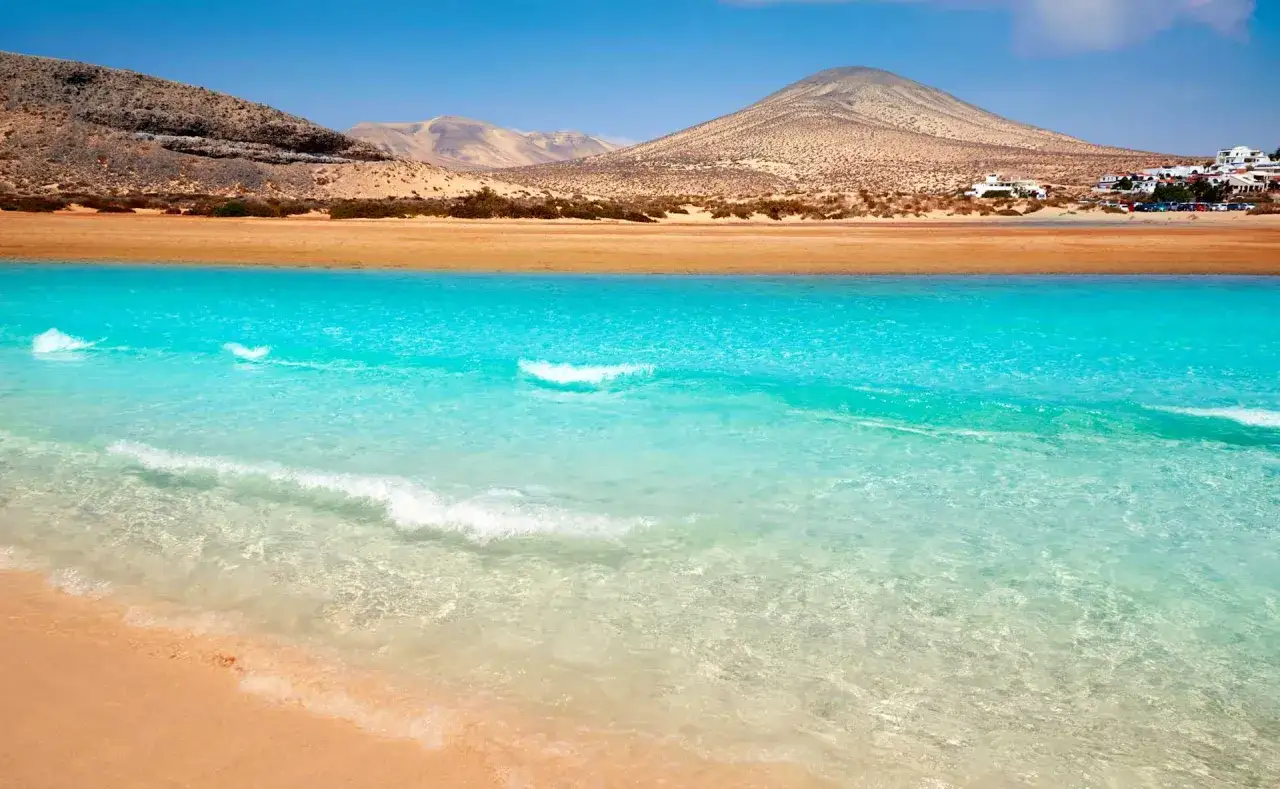 Aguas turquesas y arena dorada en las **playas Fuerteventura**, con montañas áridas y casas blancas al fondo.