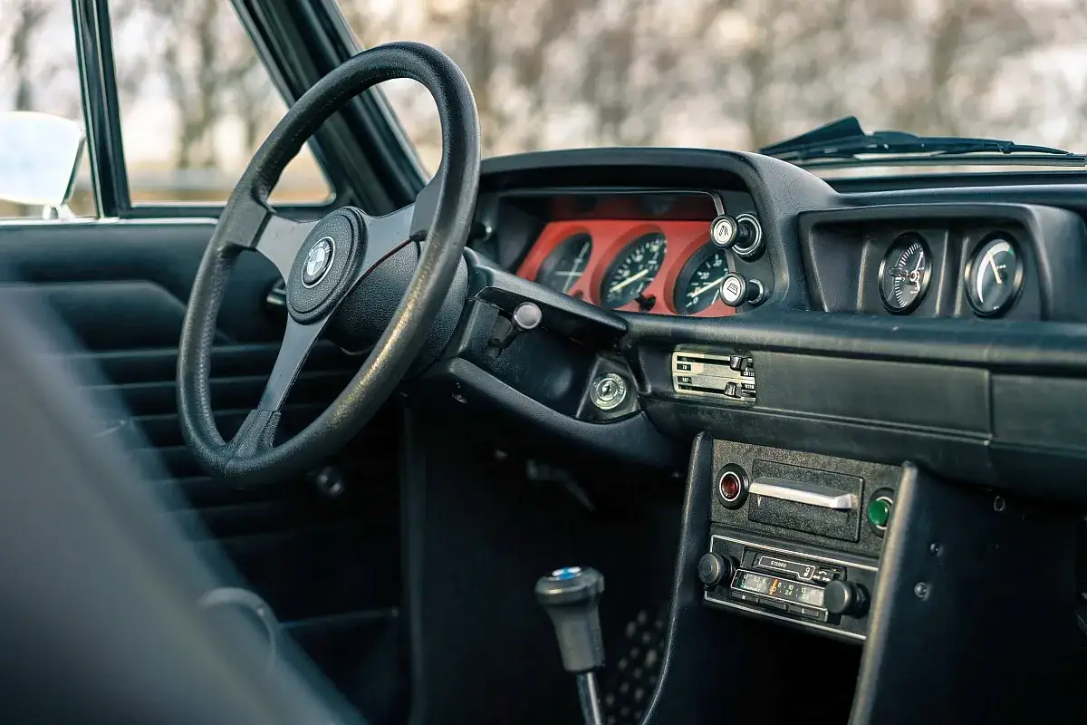 Wnętrze klasycznego BMW 2002: czarna kierownica, deska rozdzielcza z czerwonymi wskaźnikami i radiem.