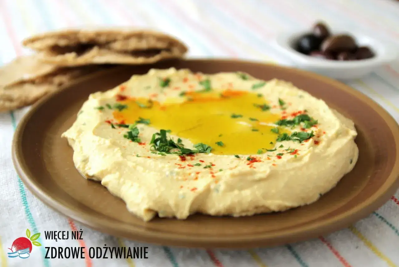 Pyszna, kremowa pasta z ciecierzycy, polana oliwą i posypana natką pietruszki i papryką. Obok pieczywo pita i oliwki.