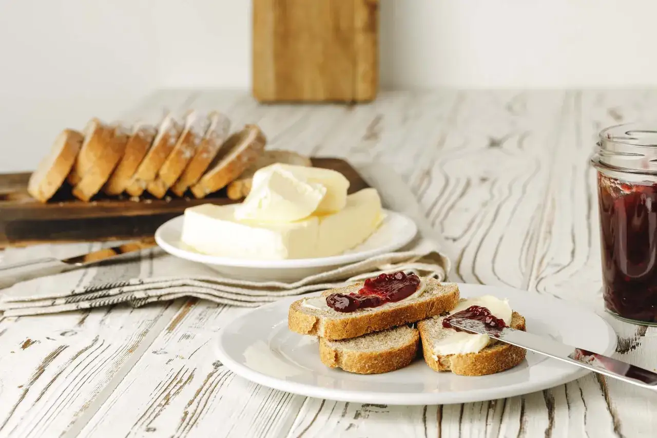 Frisches Brot mit Butter und Marmelade. Ein Glas Marmelade und ein Teller mit Scheiben von Brot, bestrichen mit Butter und Marmelade.