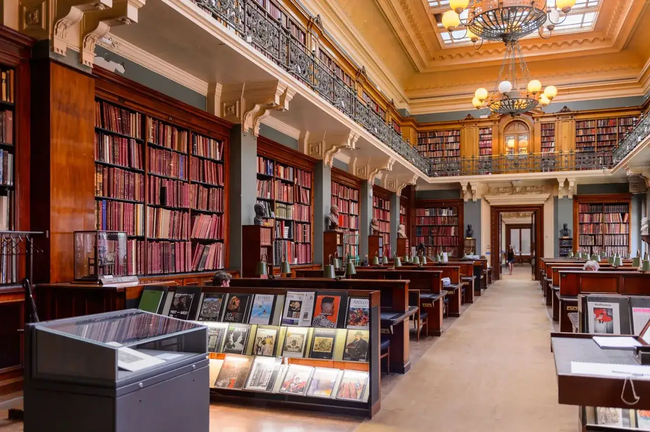 Interior de una biblioteca majestuosa, con estanterías repletas de libros que evocan **clásicos de la literatura**. Lámparas de lectura y bustos adornan los pupitres.