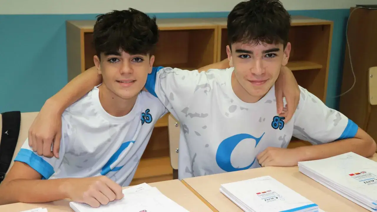 Dos chicos sonr&iacute;en, uno con el brazo alrededor del otro. Tienen camisetas blancas con un gran logo azul y el n&uacute;mero 85. Sobre la mesa, hay folletos que parecen ser una lista de material escolar.
