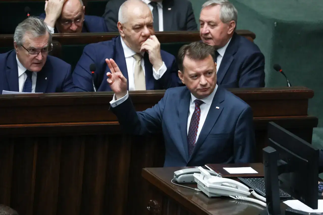 Poseł PiS przemawia na sali sejmowej, podnosząc rękę. W tle inni posłowie.