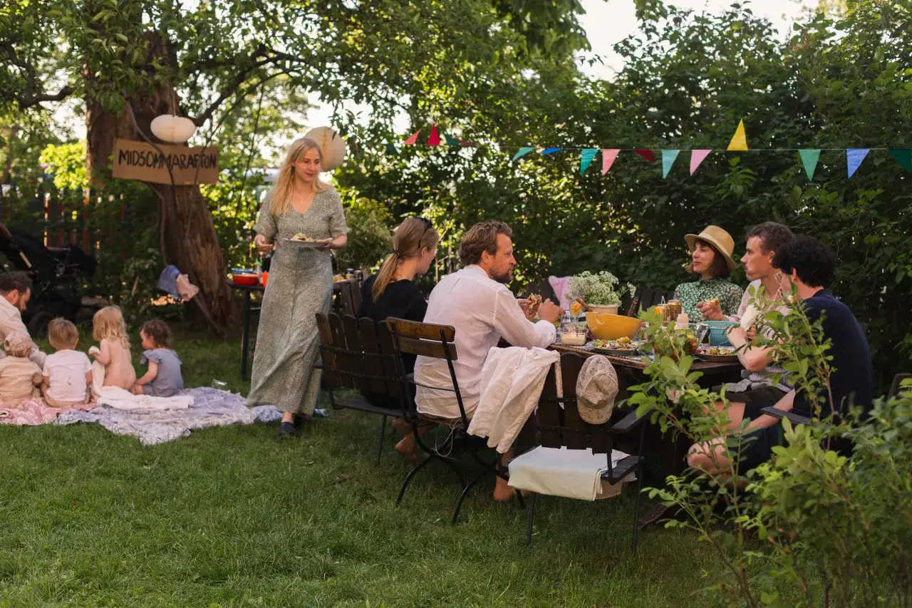 En familj firar midsommar med en härlig utomhusmåltid. Barnen sitter på filtar i gräset medan de vuxna njuter av maten vid ett dukat bord.