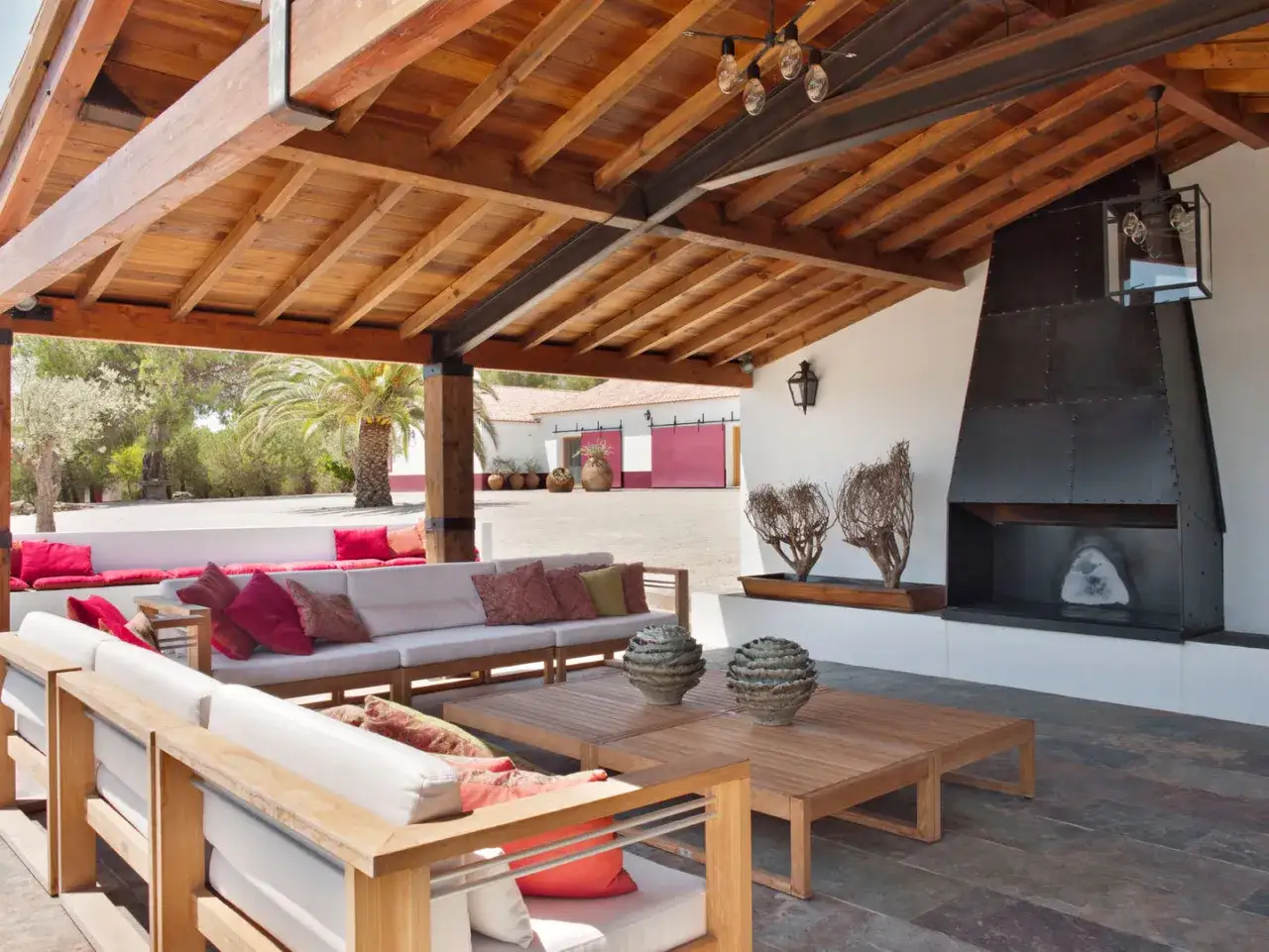 Amplia terraza con sof&aacute;s blancos, mesas de madera y chimenea met&aacute;lica, bajo **techos de madera** r&uacute;sticos.