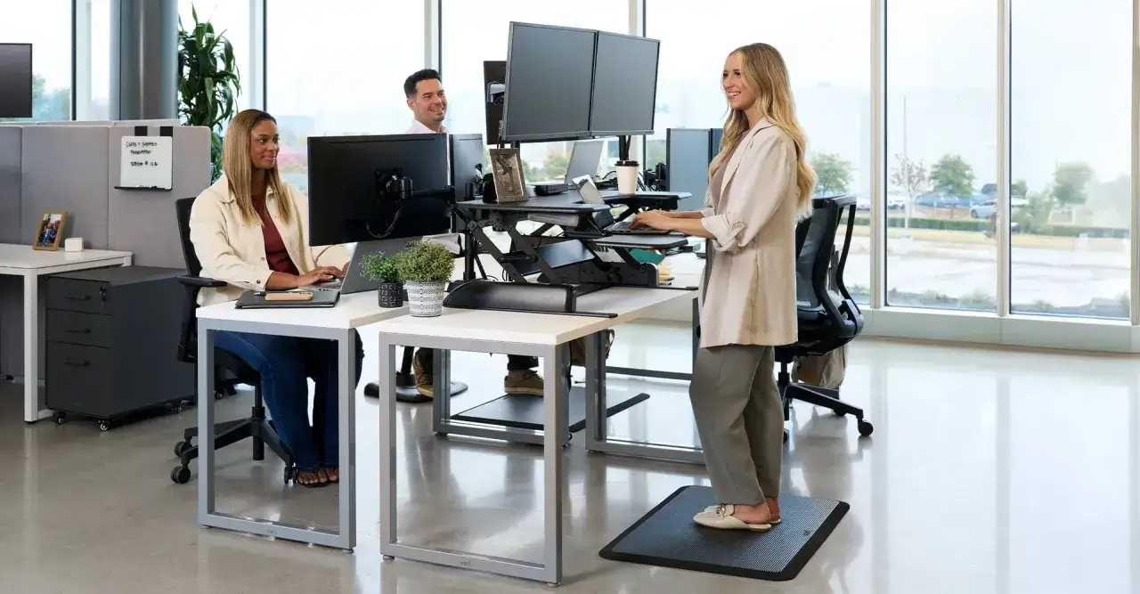 Trois coll&egrave;gues travaillent dans un bureau moderne. Une femme est assise &agrave; son bureau, tandis qu'une autre est en **position debout statique** devant son poste de travail r&eacute;glable en hauteur.