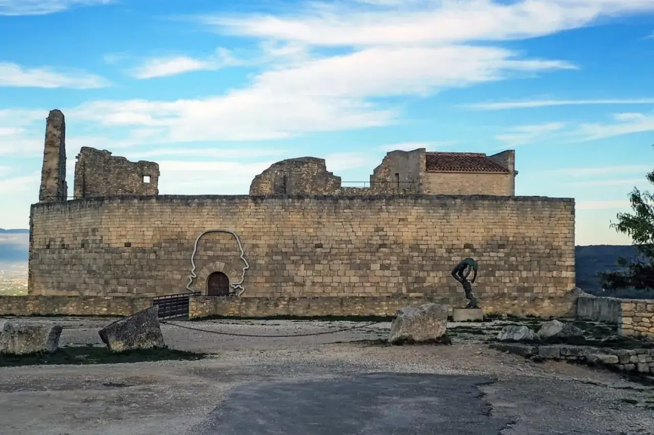 Le **ch&acirc;teau du marquis de Sade**, en ruines, se dresse sous un ciel bleu. Une sculpture de t&ecirc;te humaine orne le mur de pierre.