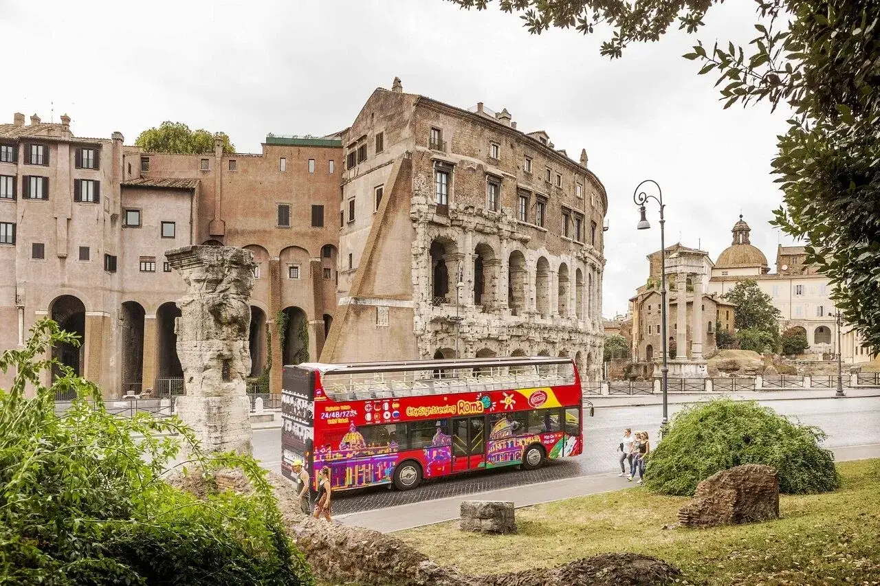 Un bus touristique rouge vif, orné d'images colorées de monuments, circule devant des ruines antiques. Un excellent moyen de faire un **rome bus tour** !