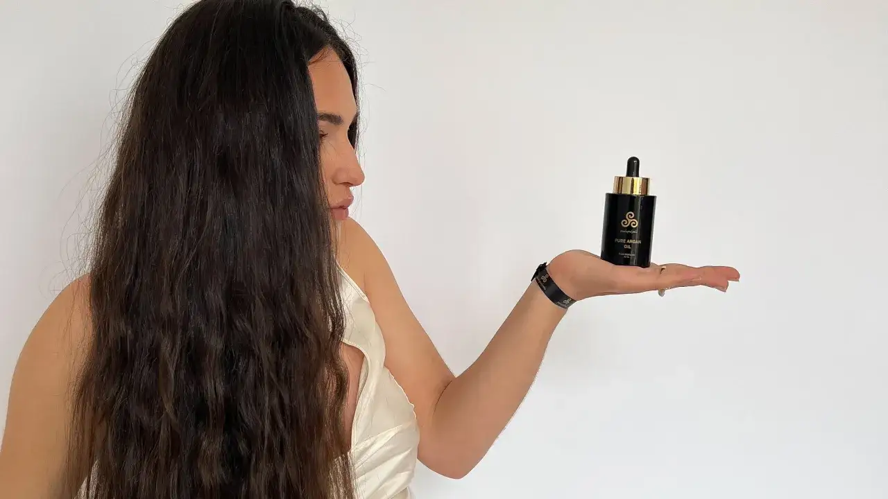 Mujer con cabello largo y oscuro sostiene un frasco de aceite de arg&aacute;n para el cabello.
