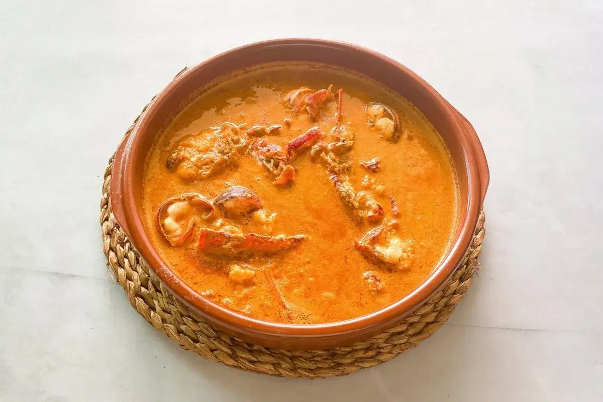 Caldereta de langosta servida en cazuela de barro sobre una esterilla de mimbre. El plato tiene un color naranja intenso y se aprecian trozos de langosta.